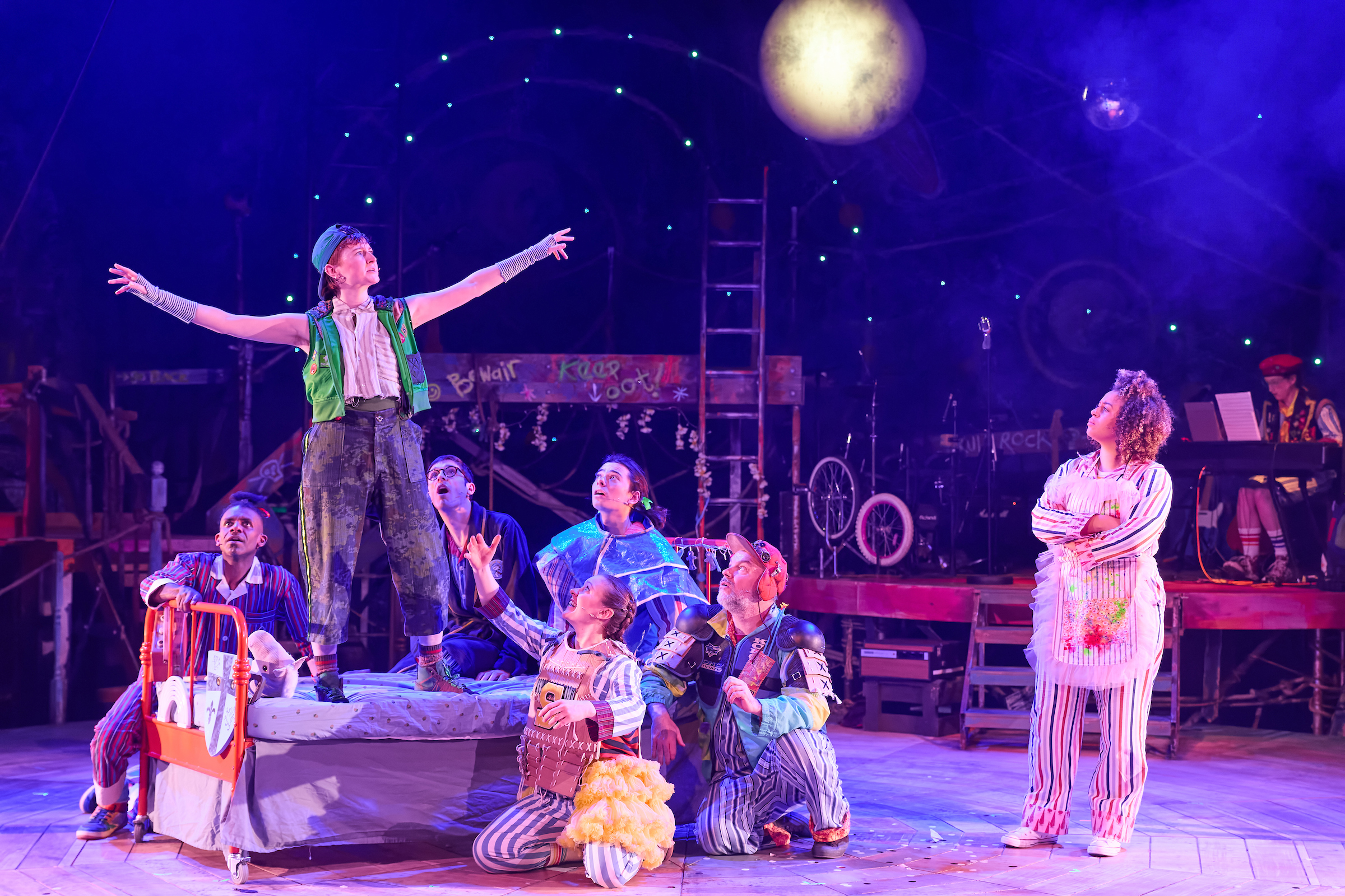 Production shot of Peter Pan at the Sherman Theatre showing the actress who plays Wendy looking at Peter Pan, standing on a bed with their arms outstretched.