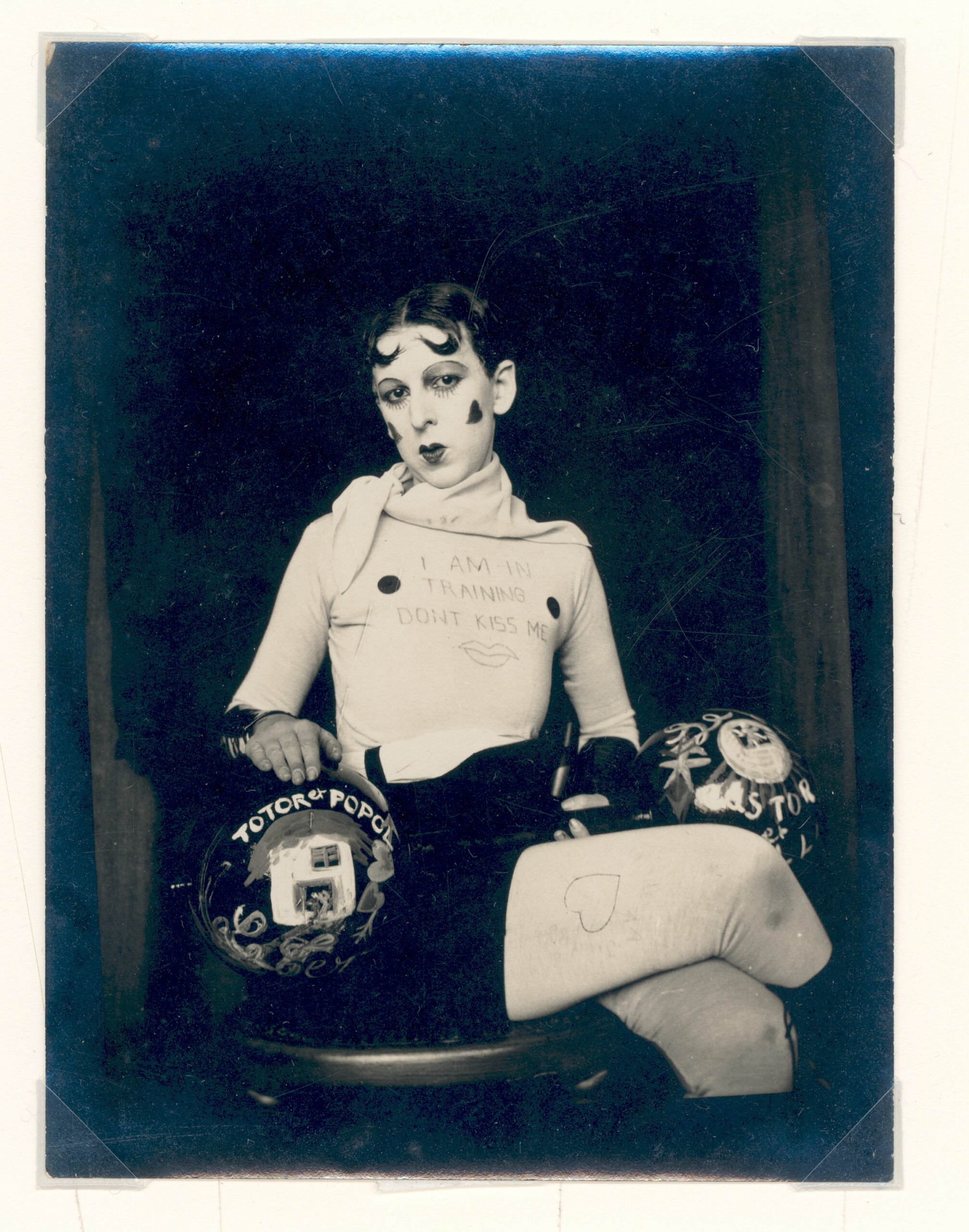A photo of Claude Cahun. Her hair and make-up is in the typical style of 1920s flapper women. Her cheeks are adorned with hearts, and her top has a message written on it beside circular nipples, proclaiming: ‘I am in training don’t kiss me.’ She has dumbbells resting on her lap so that the large balls of the weights sit on either side of her lap. This picture is presented as part of the exhibition Claude Cahun: Beneath This Mask