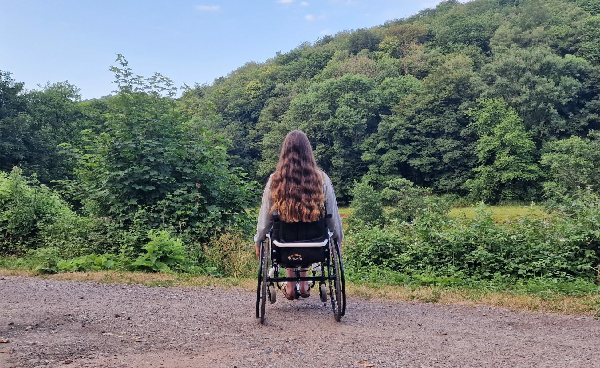 Girl with long brown hair siting in her wheelchair on a muddy pathway look into the trees