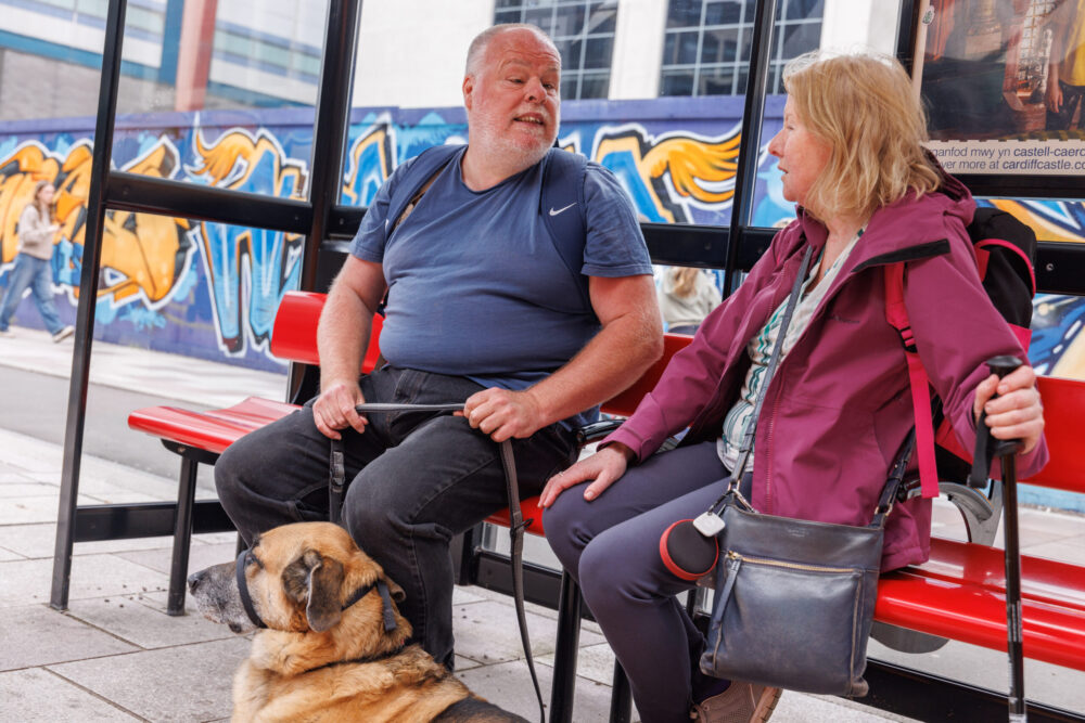 Partially sighted people with a guide dog sat at a bus stop.