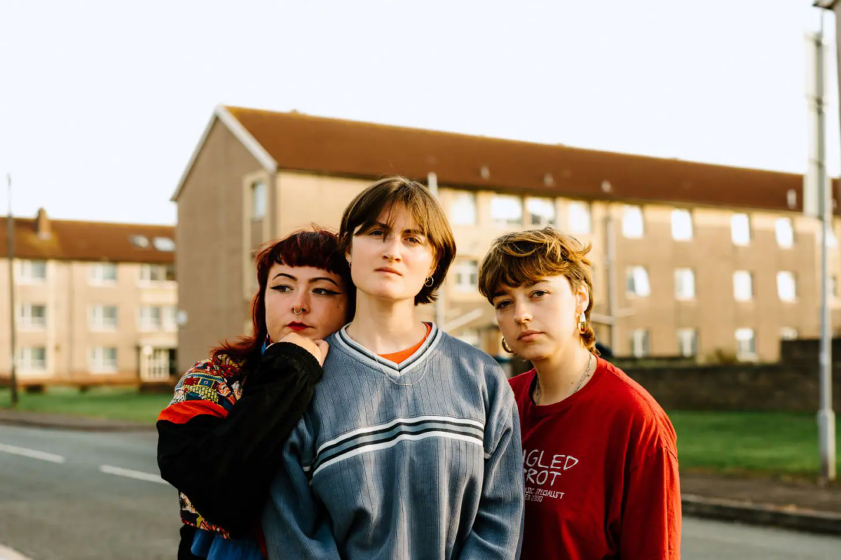 A picture of Welsh language music band Adwaith. All three band members are facing the camera in front of a house. The sun is lighting up their faces.