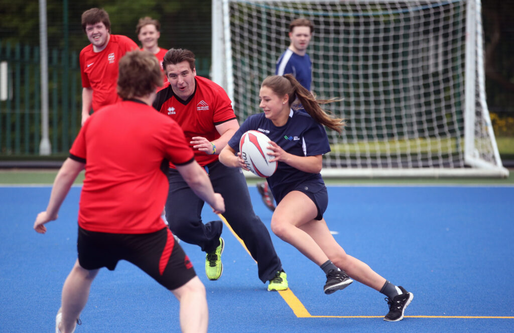 A photograph showing 6 white teenagers engaged in a game of rugby. They are playing on what appears to be a school sportsground with blue concrete, and there is a football goal net is visible in the background. The photograph is focused on two boys who seem to be about to tackle a girl, who is moving at speed and holding the ball. They all seem to be having fun.