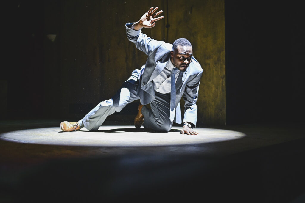 A picture of Kel Matsena, one of the cast members of Joseph K and the Cost of Living, crouching under a harsh white light