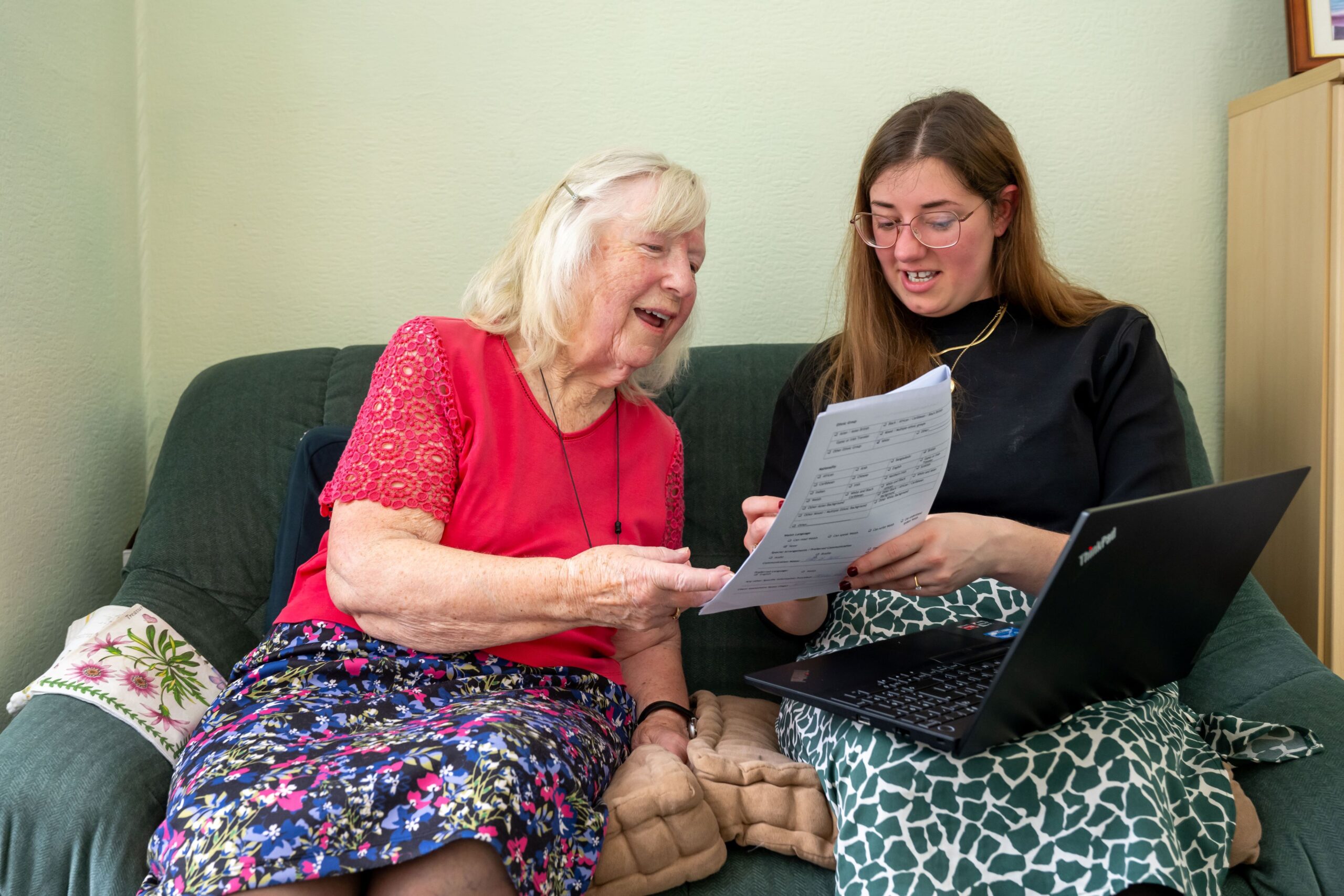 An older woman wearing a pink teeshirt and a flowered dress is speaking to a younger woman in a black top and green and white skirt. They are seated side by side on a couch and appear to be discussing a document held by the younger woman. They both seem excited and at ease. They are likely discussing Older not Colder, a new scheme to help older people keep their homes warm.