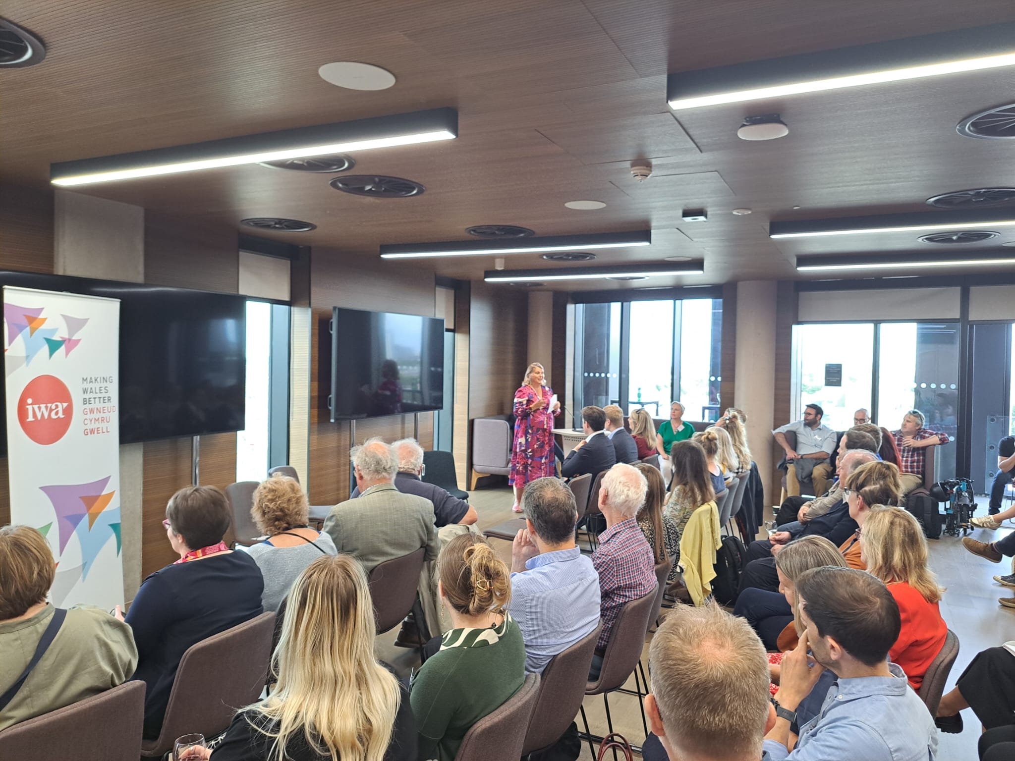 A picture of the IWA's recent summer reception. Attendees are seated and listening to Bethan Darwin, chair of the IWA. Bethan is wearing a pink and blue dress.