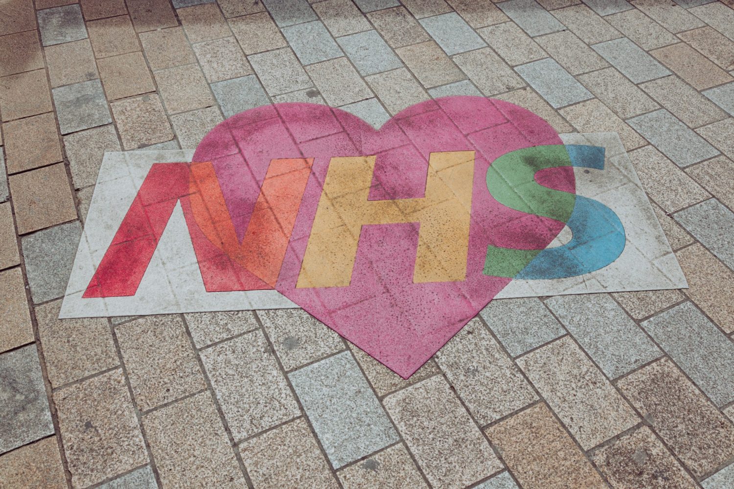A colourful NHS logo over a pink heart, painted on the ground. Photo by Nicolas J Leclercq on Unsplash.
